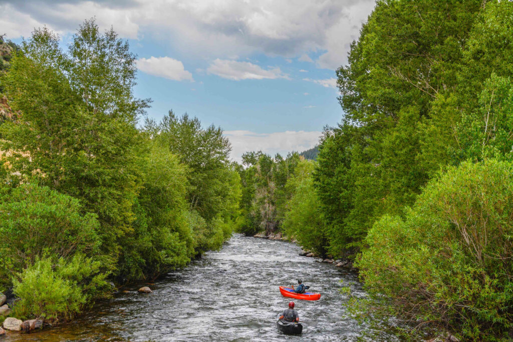 two people kayaking down river. Greenery and rocks on either side.