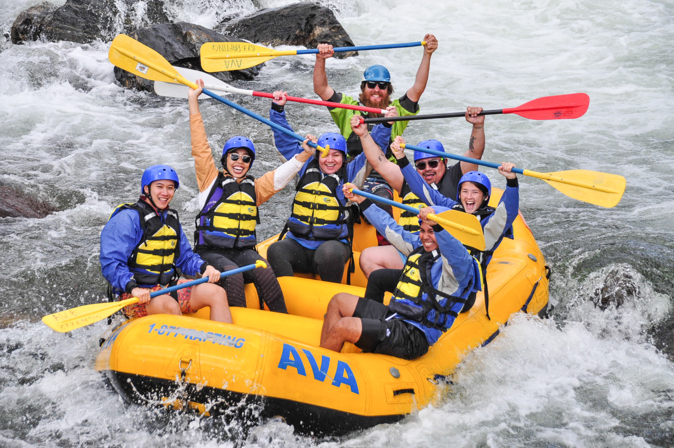 A group of people smiling with their paddles up in the air while whitewater rafting