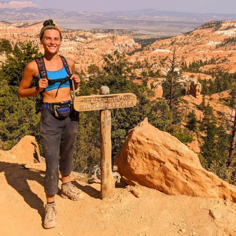 Woman standing next to wooden sign that says "Bryce Canyon Wilderness Area" with a scenic view overlooking mountains and valleys