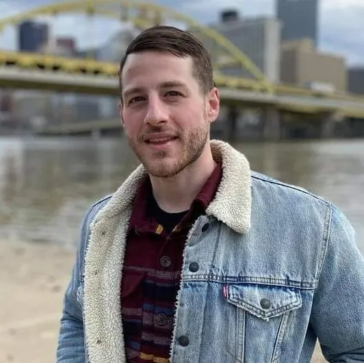 Man wearing a denim jacket on a shoreline with a skyline and yellow bridge in the background